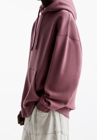 Uomo che indossa un ampio hoodie color malva con tasca a marsupio e pantaloni chiari, in piedi di lato contro uno sfondo bianco semplice.