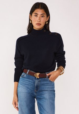 Femme portant un pull bleu marine avec des détails à boutons sur l'épaule, des boucles d'oreilles en anneau doré, une ceinture marron avec une grande boucle dorée, et un jean bleu.