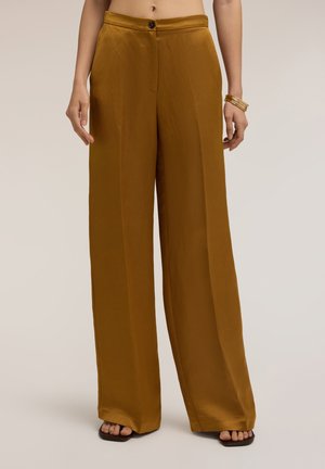Bruine satijnen broek met hoge taille en wijde pijpen met knoop aan de voorkant en plooi detail, gedragen met gouden armbanden en zwarte sandalen met open teen.