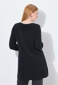 Kobieta z długimi, czerwonymi włosami, ubrana w czarną tunikę z długim rękawem i srebrnymi kolczykami w kształcie kół, stojąca przodem do białej ściany, na której widać cień.