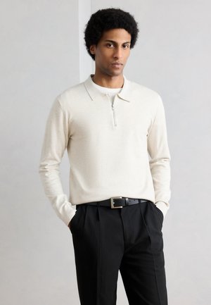 Man met krullend haar, een crèmekleurig poloshirt met lange mouwen in zwarte broek gestopt, met handen in de zakken, tegen een effen achtergrond.