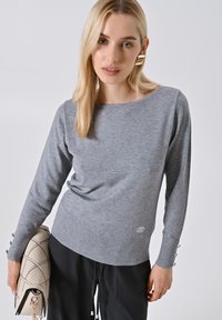 Maglione grigio lavorato a maglia con maniche lunghe, caratterizzato da bottoni decorativi sui polsini e un design fitted. Abbinato a una borsa leggera.