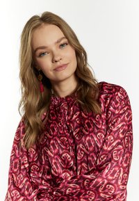 Blusa a maniche lunghe con pattern rosso e rosa, realizzata in tessuto leggero, con scollatura arricciata e orecchini con nappine decorativi.