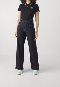 Svart polo-skjorta med guldaccenter, kombinerad med högmidjade mörka denimjeans. Modellen bär vita sneakers med gröna detaljer.