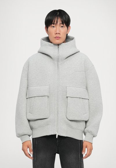Haut à zip gris avec de grandes poches avant, col haut et tissu texturé. Associé à un jean foncé, à la coupe décontractée.