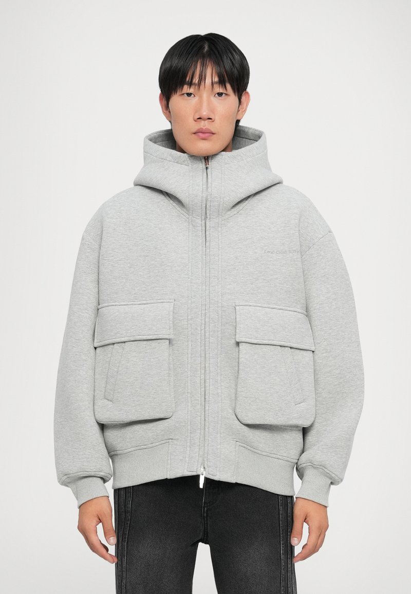 Haut à zip gris avec de grandes poches avant, col haut et tissu texturé. Associé à un jean foncé, à la coupe décontractée.
