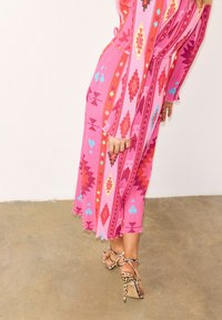 Γυναίκα που φοράει ροζ midi φόρεμα με σχέδια και ψηλοτάκουνα σανδάλια με λεοπάρ σχέδιο, στέκεται σε τσιμεντένιο πάτωμα μπροστά σε λευκό τοίχο.