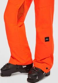 Heldere oranje waterdichte skibroek met gaasventilatie, een zwart label op het onderbeen en zwarte skischolen met verstelbare gespen.
