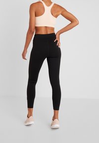 Osoba w jasnoróżowym staniku sportowym, czarnych legginsach i jasnoróżowych butach sportowych stoi z jedną ręką na biodrze na tle jednolitej szarej ściany.