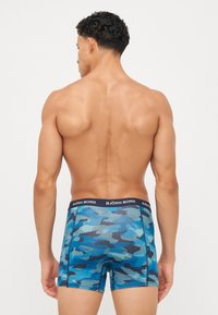 Blaue Camouflage-Boxershorts mit einem schwarzen Bund, auf dem der Markenname "BJÖRN BORG" steht. Hergestellt aus dehnbarem Material und in einer Länge über dem Knie.