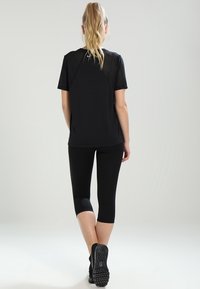 Mujer con coleta rubia de pie, de espaldas a la cámara, vistiendo una camiseta negra de manga corta, leggings capri negros y zapatillas negras.