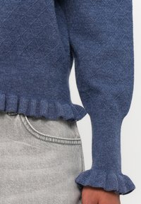 Suéter azul com um padrão de diamante texturizado, punhos canelados e uma bainha em renda. Combinado com calças de ganga cinzas, apresentando um ajuste descontraído.