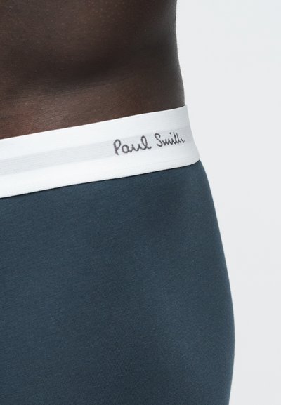 Gros plan sur la taille d'une personne portant des sous-vêtements bleu foncé avec une ceinture blanche portant l'étiquette "Paul Smith."