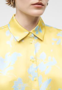 Κοντινό πλάνο ενός ατόμου που φοράει κίτρινο πουκάμισο με κουμπιά, διακοσμημένο με φωτεινά μπλε λουλουδένια σχέδια και χρυσούς κρίκους σκουλαρίκια.
