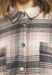 Camicia di flanella a quadri con colletto; presenta quadri rosa, neri e bianchi. Include bottoni e una tasca per un ulteriore dettaglio.