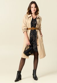 Beiger Trenchcoat mit Gürtel, schwarzes Kleid mit floralem Muster, schwarze Handtasche, blickdichte Strumpfhosen und schwarze Stiefeletten mit Blockabsatz.