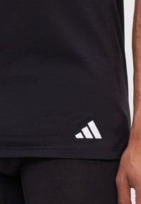 Juodas sportinis marškinėlis, pagamintas iš medvilnės, su lygiu apvadu ir baltu trimis juostelėmis paženklintu logotipu kairėje pusėje.