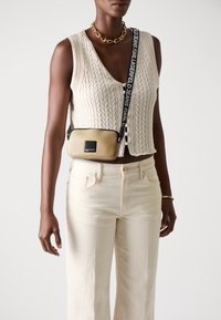Beige synthetische Umhängetasche mit einem schwarzen Logo-Patch. Merkmale einen gemusterten Riemen mit Text und eine glatte Oberfläche. Wird über einem Strickwesten getragen.