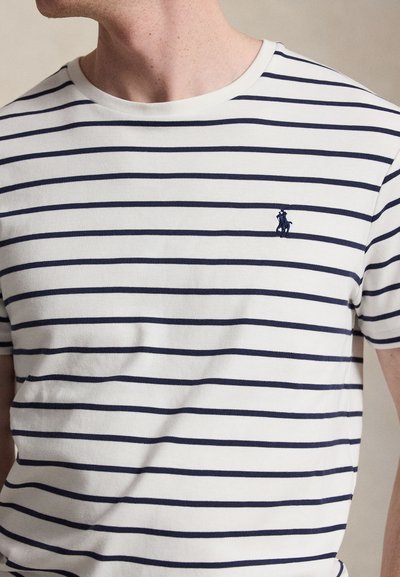 Λευκό μπλουζάκι με οριζόντιες ναυτικές ρίγες και μικρό ναυτικό κεντητό λογότυπο στο αριστερό στήθος, φορεμένο από ένα άτομο που κοιτάζει ελαφρώς πλάγια.