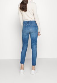 Jeans skinny de tiro alto en azul, hechos de denim, con dos bolsillos traseros y una silueta ajustada. Combinados con zapatillas blancas.