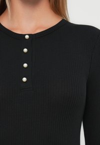 Top lung cu maneci lungi, texturat, de culoare neagră, cu guler rotund și o închidere cu cinci nasturi din perle. Materialul este moale și se potriveste bine.