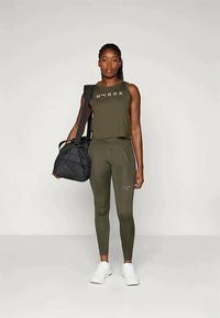 Olive green athletic tank top with "HYROX" text, paired with matching leggings. White sneakers and a black gym bag complete the outfit.
