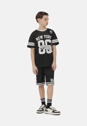 Ragazzo in abbigliamento sportivo nero New York 86 con una grafica di tigre, pantaloni corti abbinati, calze a righe e sneakers in bianco e nero, che guarda di lato.
