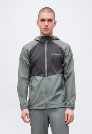 Jeune homme debout face à l'avant, portant une veste à capuche zippée Columbia verte et gris foncé et un pantalon assorti.