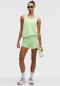 Canotta sportiva verde chiaro e pantaloni corti realizzati in materiale traspirante. Gli accessori includono occhiali da sole con lenti gialle e una bottiglia d'acqua trasparente.