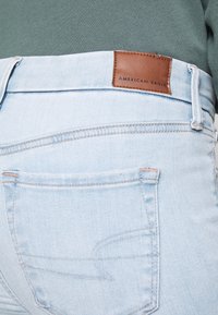 Los jeans de mezclilla azul claro cuentan con una etiqueta de cuero marrón con texto en relieve, costuras de contraste sutil y diseños de bolsillos traseros angulados.