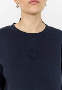 Navy sweatshirt with ribbed crew neck, featuring an embossed circular logo on the chest. Soft texture, long sleeves, casual fit.