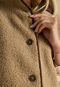 Beige Fleecejacke mit strukturierter Oberfläche, braunen Besätzen entlang der Kanten und einem runden Knopfverschluss mit eingraviertem Text.