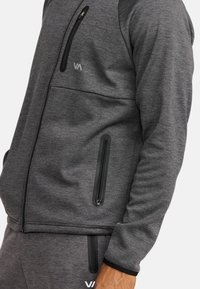 Veste gris zippée en tissu texturé, avec des poches zippées noires, des poignets côtelés et un petit logo sur la poitrine.