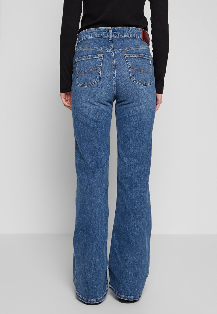 Un jean en denim bleu évasé présente une taille haute, deux poches arrière avec des détails cousus et une étiquette en cuir à la taille.
