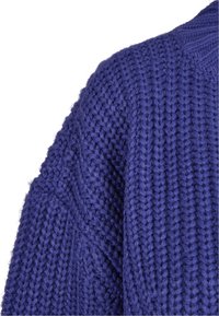 Gros plan sur une épaule de pull bleu tricoté montrant un fil épais et texturé avec un motif côtelé sur fond blanc.
