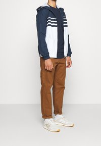 Veste légère colorée en bleu marine et blanc avec capuche à cordon, poignets élastiques et trois bandes blanches sur le torse, accompagnée de pantalons marron et de baskets.