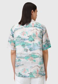Camisa de manga corta de tejido ligero que presenta un patrón de paisaje tropical con palmeras, montañas y flores coloridas.
