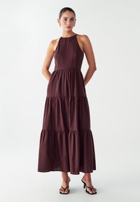 Nainen, jolla on yllään hihaton tummanruskea kerroksellinen maksimekko, jossa on korkea pääntie, sekä mustat korkokengät, seisoo valkoista taustaa vasten.