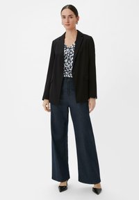 Zwarte blazer met een getailleerde pasvorm over een bedrukte top, gecombineerd met wijde donkerblauwe denim broek en puntige zwarte hakken. Simpele accessoires.