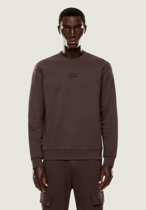 Mand iført brun langærmet sweatshirt og matchende cargobukser med EA7-logo, stående mod en ensartet lys baggrund.