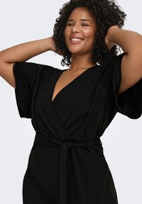 Juodas kombinezonas su trumpomis plačiomis rankovėmis, gilus V formos iškirptė ir surišama liemens juosta. Minkšta medžiaga su lygiu paviršiumi.