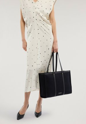 Mujer con vestido blanco sin mangas de lunares, sosteniendo un bolso grande negro, lleva tacones negros de punta afilada, de pie sobre un fondo blanco.