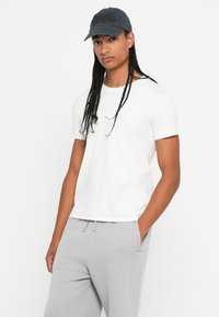 Hvid bomulds T-shirt sat sammen med grå sweatpants. Kasketten er mørkegrå med et struktureret logo foran. Modellen står i en afslappet positur.