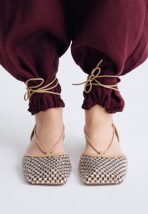 Pies con zapatos planos beige cubiertos de pedrería, combinados con pantalones granate atados en los tobillos con cordones beige.