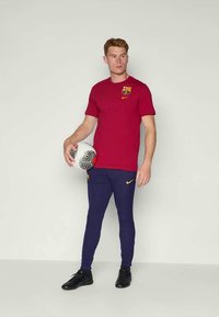 Červené tričko s krátkym rukávom Nike s logom Barcelony a žltým akcentom, v kombinácii s námorníckymi rebrovanými atletickými nohavicami a čiernymi topánkami, drží futbalovú loptu.