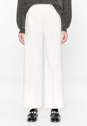 Person wearing high-waisted wide-leg white pants, dark gray sweater, and black loafers with silver embellishments, standing against a white background.