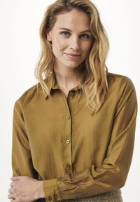 Lächelnde Frau mit welligem blonden Haar trägt eine braune, knöpfbare Bluse und schlägt die Arme vor einem einfarbigen hellen Hintergrund überkreuz.