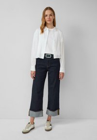 Weißes Hemd mit Knöpfen, schlichtes weißes T-Shirt, dunkle Jeans mit weitem Bein und umgeschlagenen Säumen sowie beige Sneakers mit schwarzen Akzenten.