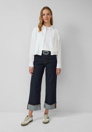 Camicia bianca con bottoni, t-shirt bianca semplice, pantaloni in denim scuro a gamba larga con orli risvoltati e sneakers beige con dettagli neri.
