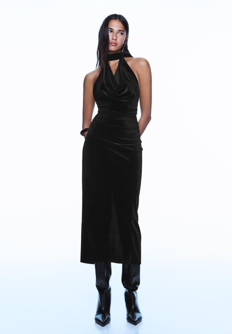 Czarna aksamitna sukienka na szyję z dekoltem typu serek, dopasowaną talią i spódnicą o długości do połowy łydki. W połączeniu z czarnymi kozakami za kolano o spiczastych noskach.